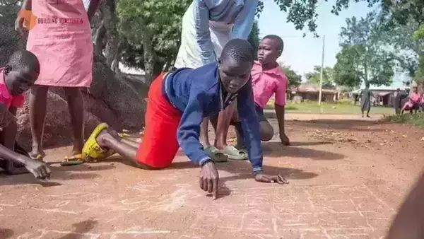 A young student, wearing a blue sweater and red skirt, kneels on the ground and traces letters and numbers written in chalk. Other children in pink shirts look on. In the background, other children play in a dusty area, and small buildings are visible. A partial logo in the upper left corner reads "Building Tomorrow."