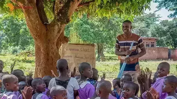 A teacher smiles at a group of enthusiastic children clapping beneath the shade of a large tree. He holds a pointer toward an expansion chart propped against the tree trunk, illustrating the concept of thousands, hundreds, tens, and ones. A small building is visible in the background.