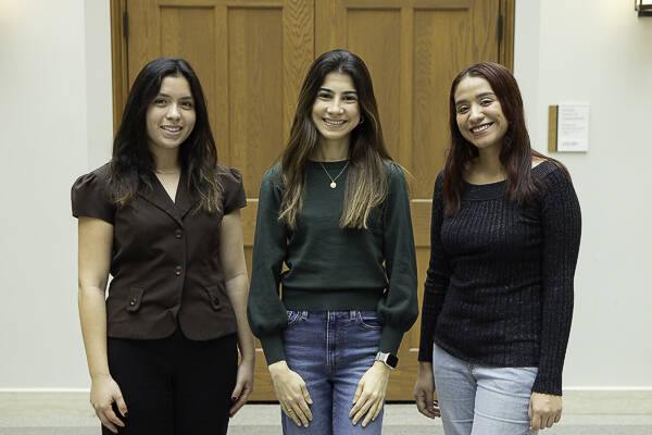 Three young women with shoulder-length brown hair smile at in front of a wooden door in a bright hallway.
